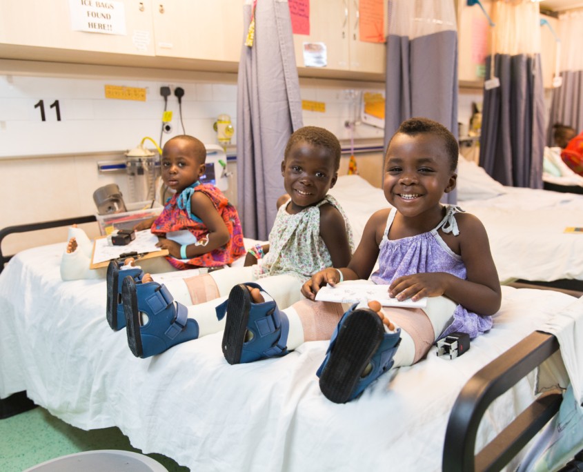 Three little orthopedic patients laugh and play on one of the ward beds.